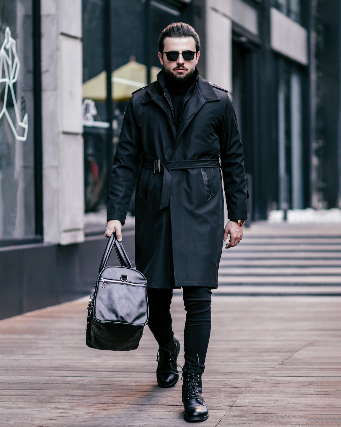 Man in black trench coat walks on wooden city walkway.