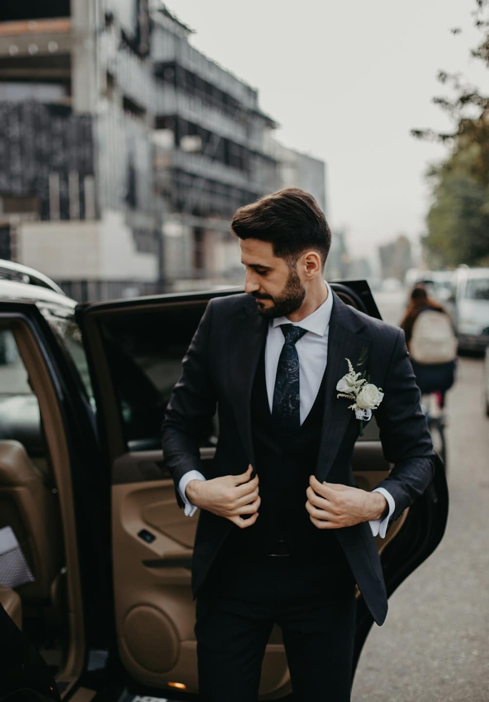 Man in suit adjusts his jacket by a car door.
